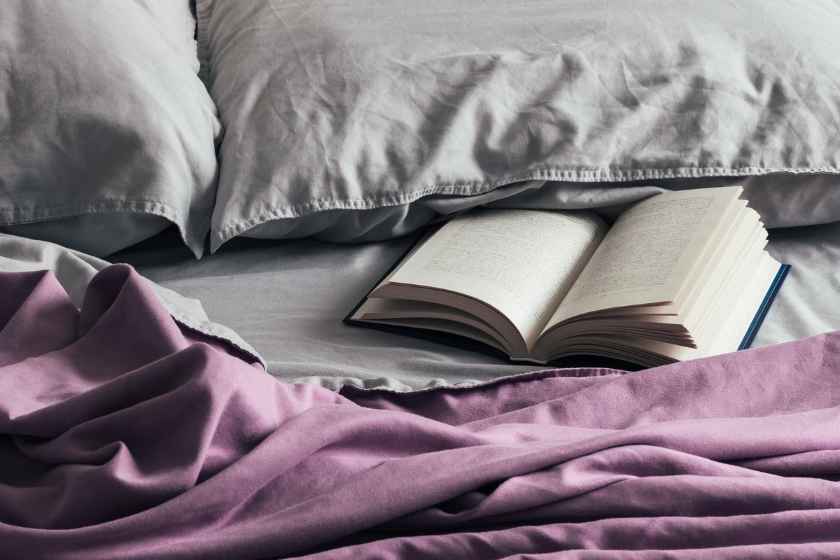Open book resting on a bed with grey pillows and a lavender blanket, a small self-care ritual before sleep