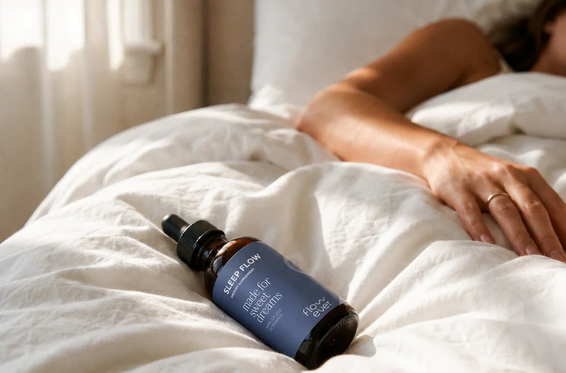 Woman sleeping under white linen sheets in soft morning light, with Sleep Flow drops resting on the bed